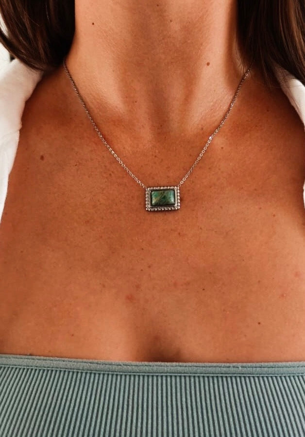 brunette model wearing a white shirt, blue top and silver pendant necklace with green stone.